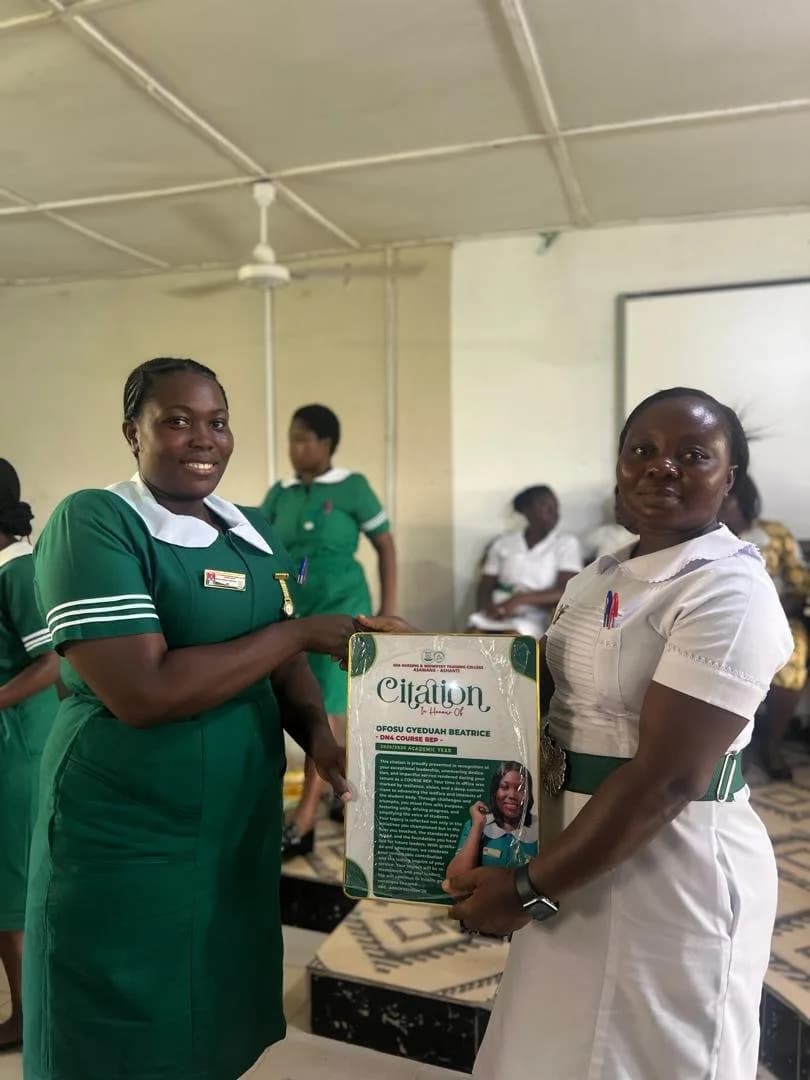 Female student in green nursing uniform presenting citation award to woman in beige dress during handover ceremony